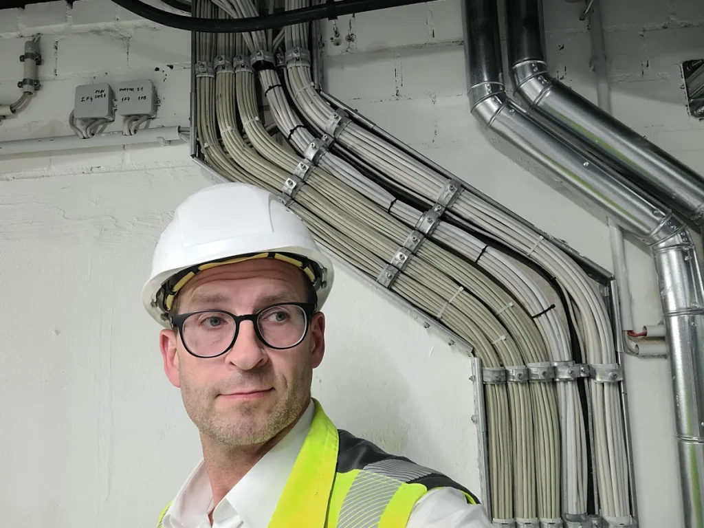 Eine Person mit weißem Schutzhelm und Sicherheitsweste steht neben einem Geflecht aus Kabeln und Rohren vor einer weißen Wand.