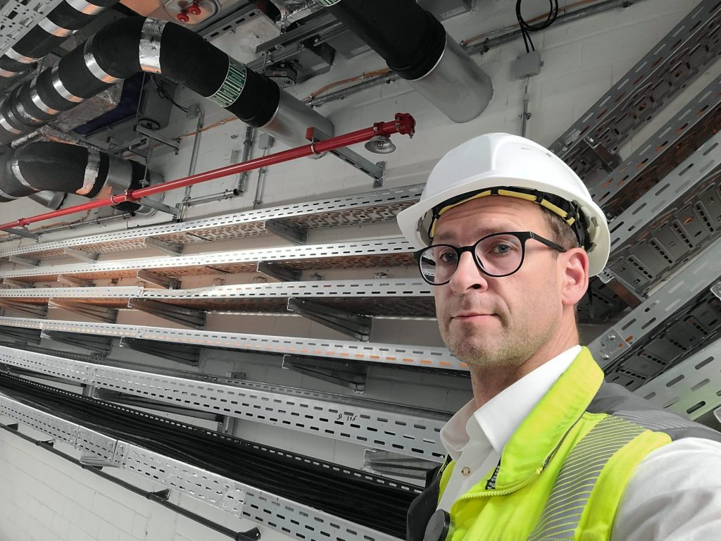 Eine Person, die einen Schutzhelm und eine Warnweste trägt, steht in der Nähe von Metallleitungen und -rohren auf einer Baustelle.