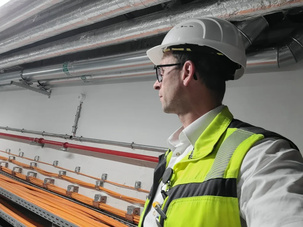 Eine Person mit Schutzhelm und Warnweste steht in einem Industrieraum, an dessen Wand Rohre und orangefarbene Kabel anliegen.