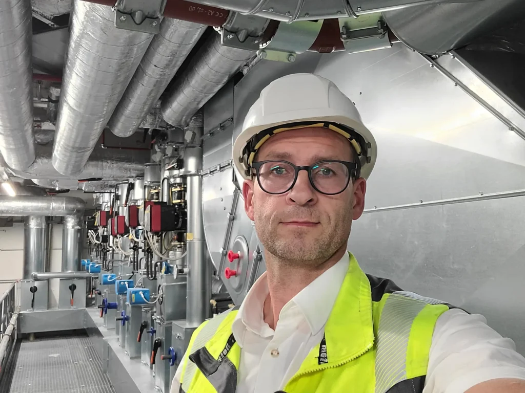 Eine Person mit Schutzhelm und Warnweste posiert in einer Industrieanlage voller Rohre und Maschinen.