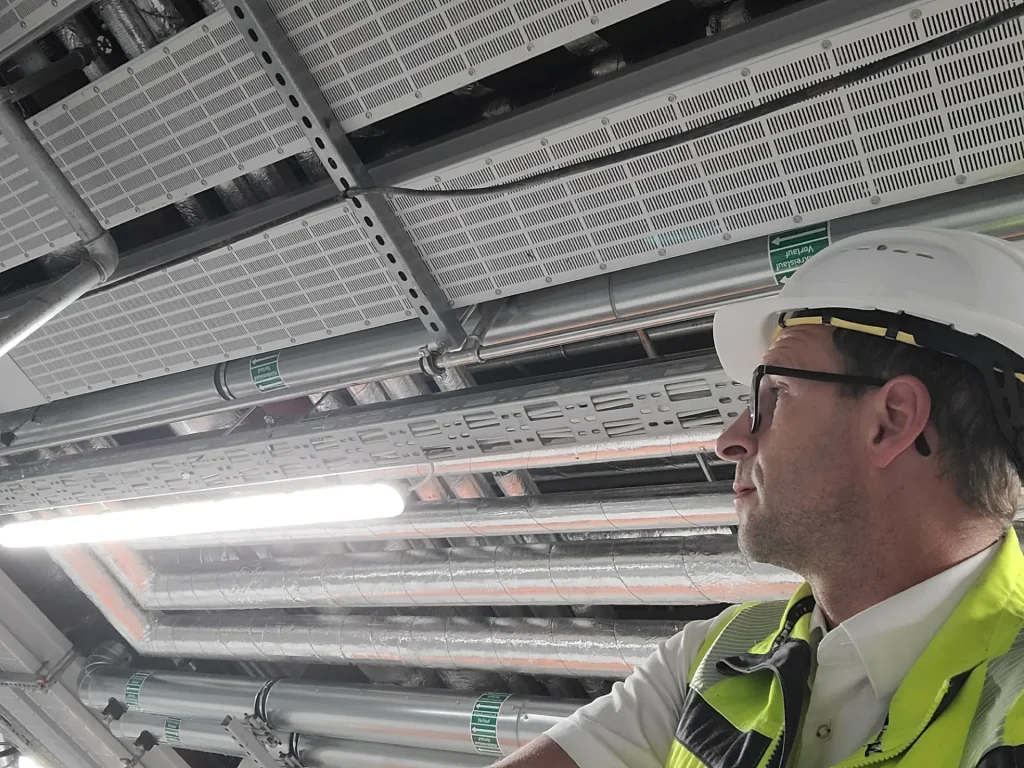 Eine Person mit Schutzhelm und Warnweste beobachtet industrielle Rohrleitungen und Beleuchtungsanlagen in einer Baustellenumgebung.