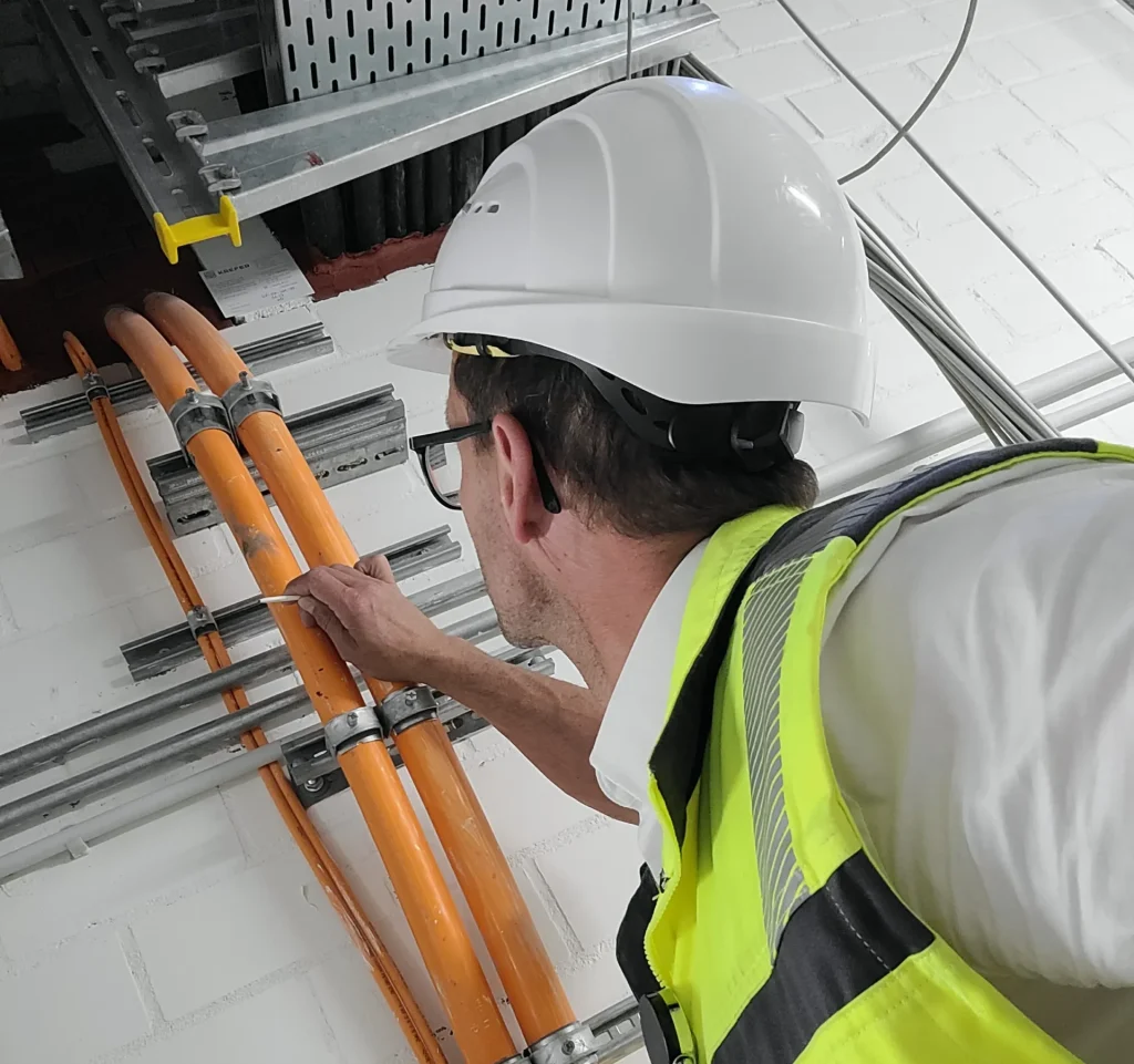 Ein Arbeiter in weißem Helm und gelber Sicherheitsweste inspiziert orangefarbene elektrische Leitungen an einer Wand, die von Metallhalterungen umgeben sind.