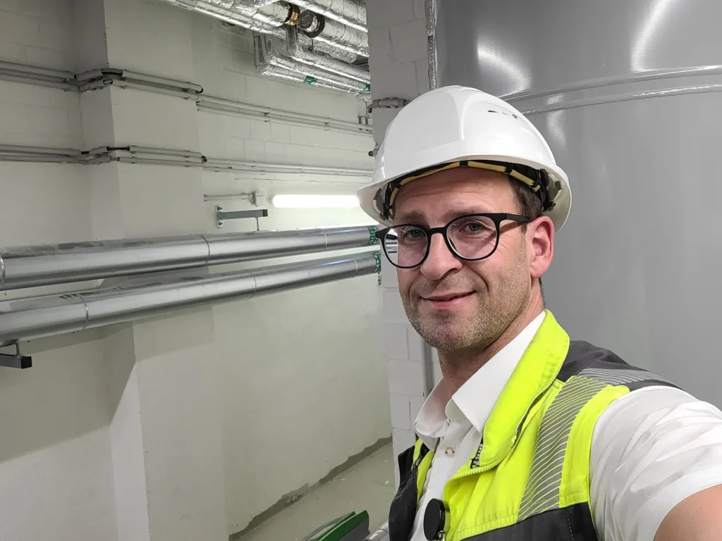 Eine Person mit Schutzhelm und Warnweste posiert für ein Selfie in einer gut beleuchteten Industrieanlage mit sichtbaren Metallrohren.
